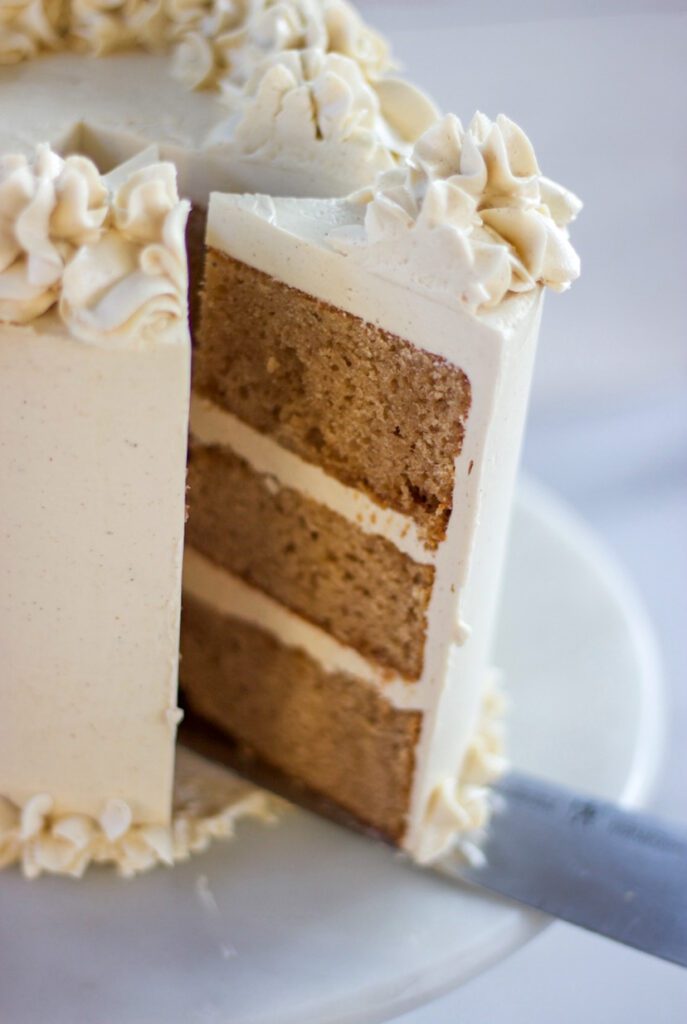 Chai Cake with white frosting and a white wood background. A slice being taken off.