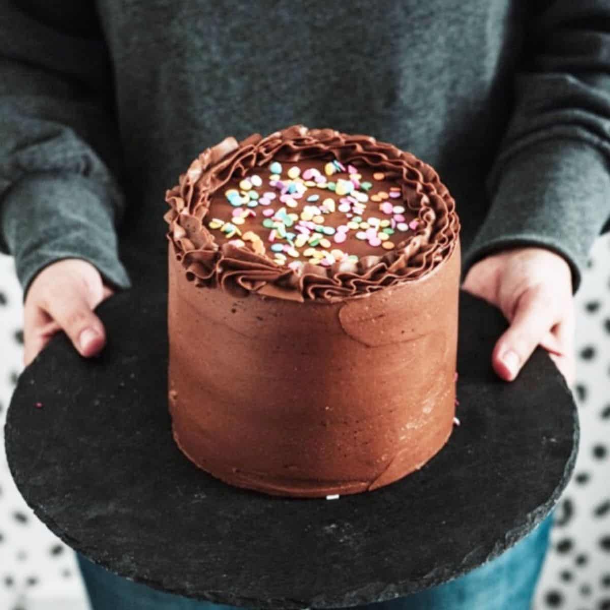 holding a 6'' chocolate cake on a board in front of body
